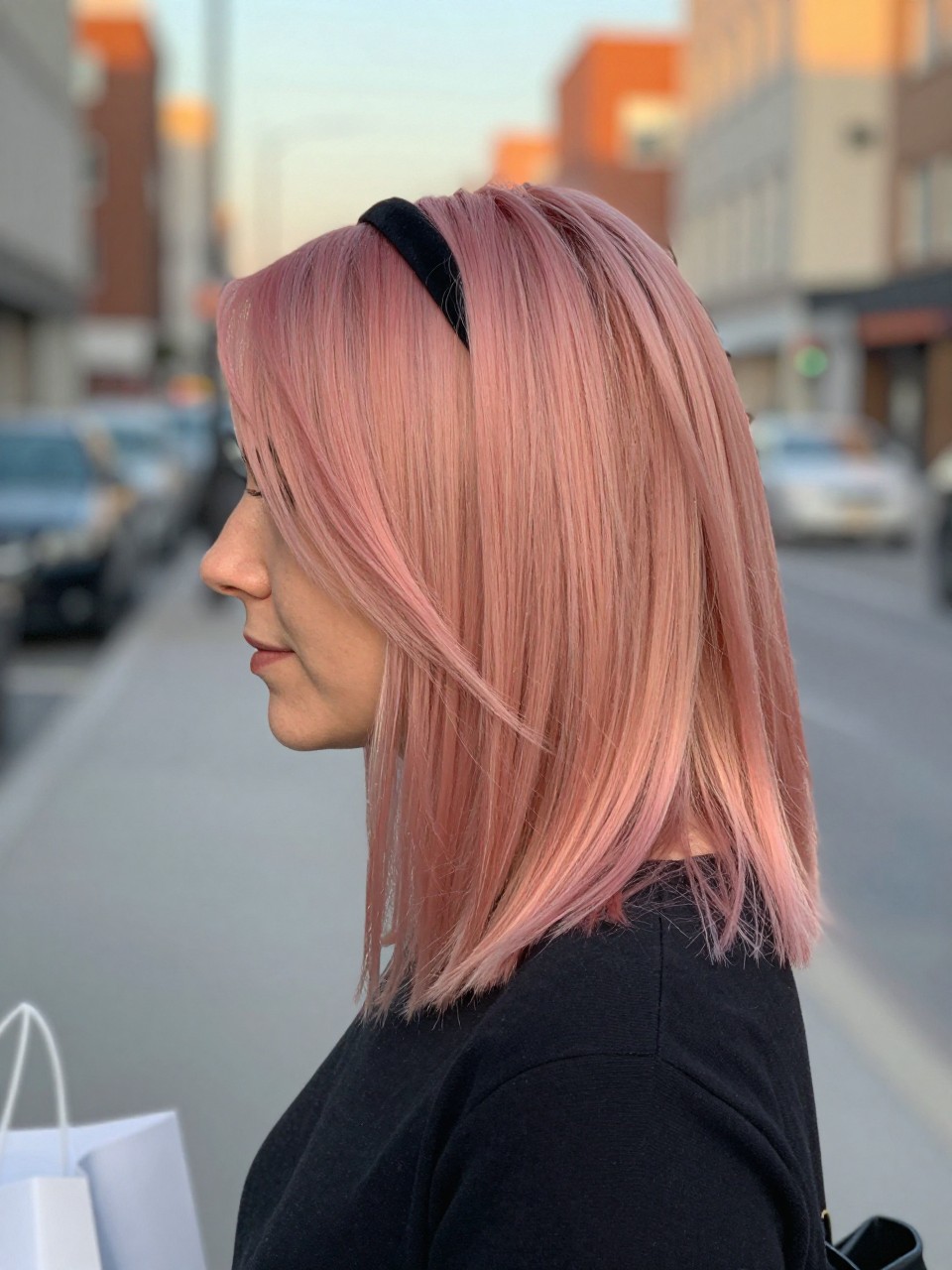 Photo of a woman wearing Blunt Cut With Soft Strawberry Melt highlights and soft texture, side profile view, in a softly blurred city street background, golden hour glow, containing a small lived-in detail such as a headband pushed back and a few shopping bags nearby, latest iPhone photo quality.