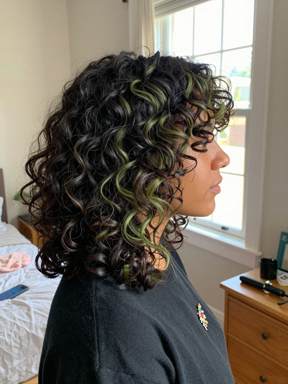 Photo of a biracial woman wearing soft black curls with olive green highlight streaks, side profile view, in a sun-drenched bedroom, natural window light, containing a small lived-in detail such as a decorative pin on one side and a curling iron resting on the nightstand, latest iPhone photo quality.