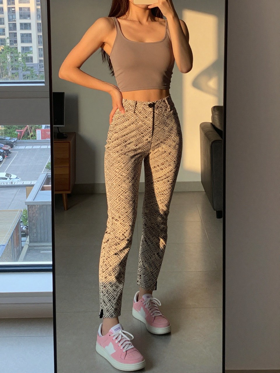 iPhone mirror selfie of a woman in printed pants and a fitted top with pink sneakers, standing with hand on hip, cropped at NECK so face doesn't show, in a city apartment, evening golden hour, iPhone photo quality.
