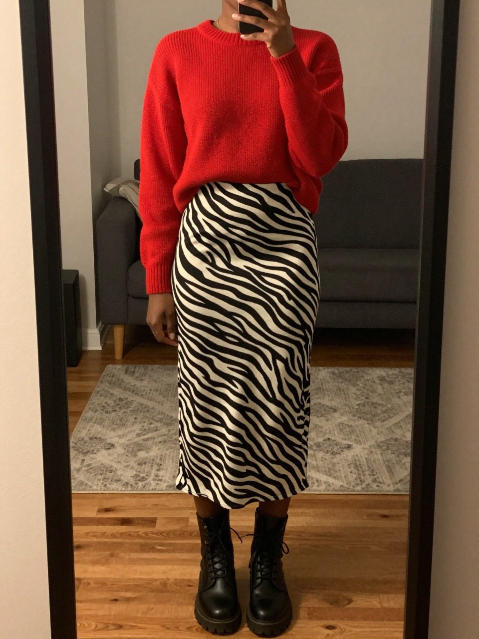 mirror selfie of a black woman in a red sweater and a zebra print long skirt with combat boots, standing with feet shoulder-width apart, cropped at NECK so face doesn't show, in a city apartment, warm moody lighting, iPhone photo quality.