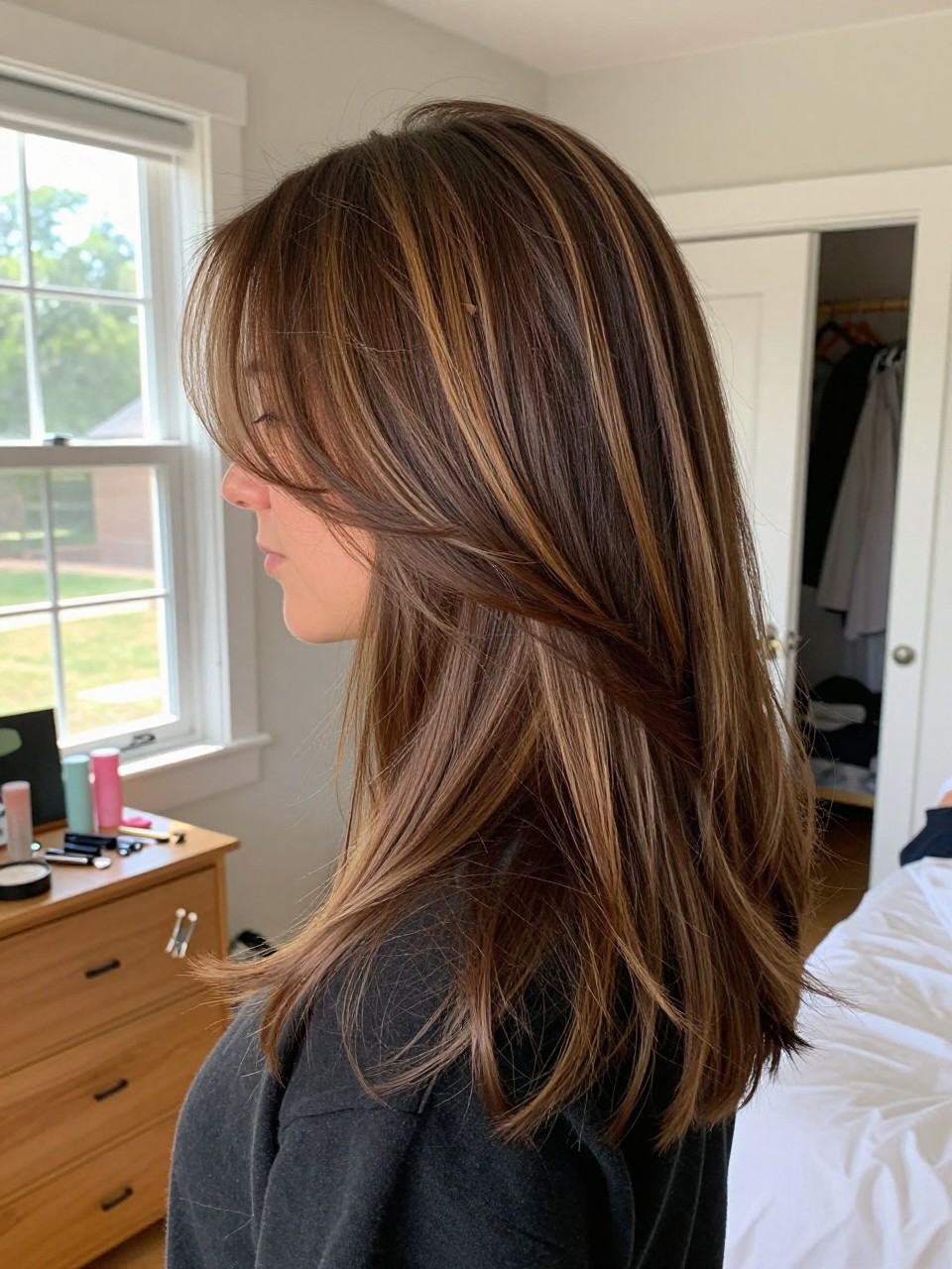 Photo of a woman wearing layered chocolate brown hair with toffee highlights and blended lowlights, side profile view, in a sun-drenched bedroom, natural window light, containing a small lived-in detail such as small sectioning clips on the dresser and a slightly open closet behind, latest iPhone photo quality.
