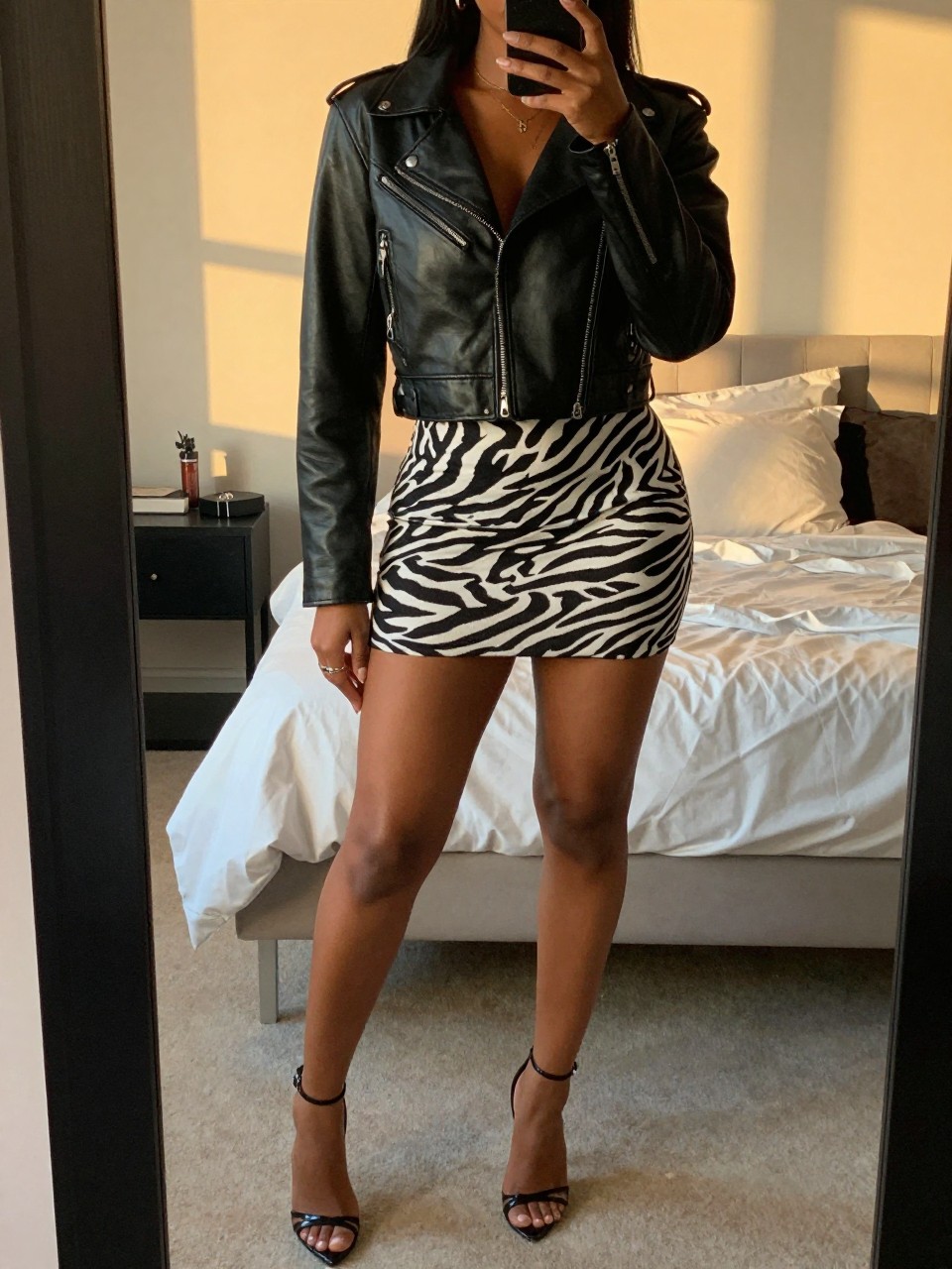 mirror selfie of a black woman in a cropped black leather jacket and a zebra print mini skirt with strappy stilettos, one hand in her jacket pocket, cropped at NECK so face doesn't show, in a feminine-themed bedroom, evening golden hour, iPhone photo quality.