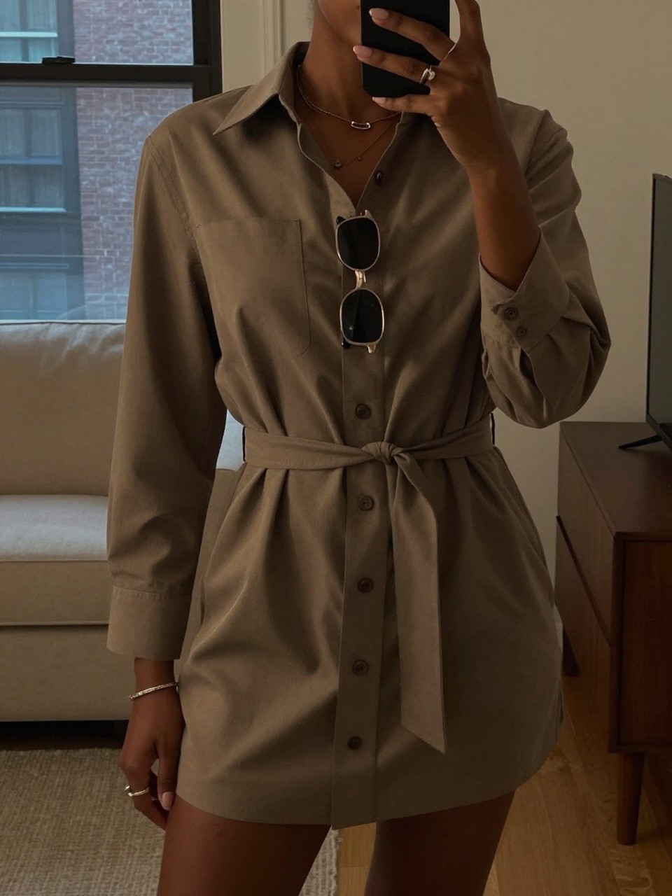mirror selfie of a black woman in a belted shirt dress, holding sunglasses, cropped at NECK so face doesn't show, in a city apartment, warm moody lighting, iPhone photo quality.