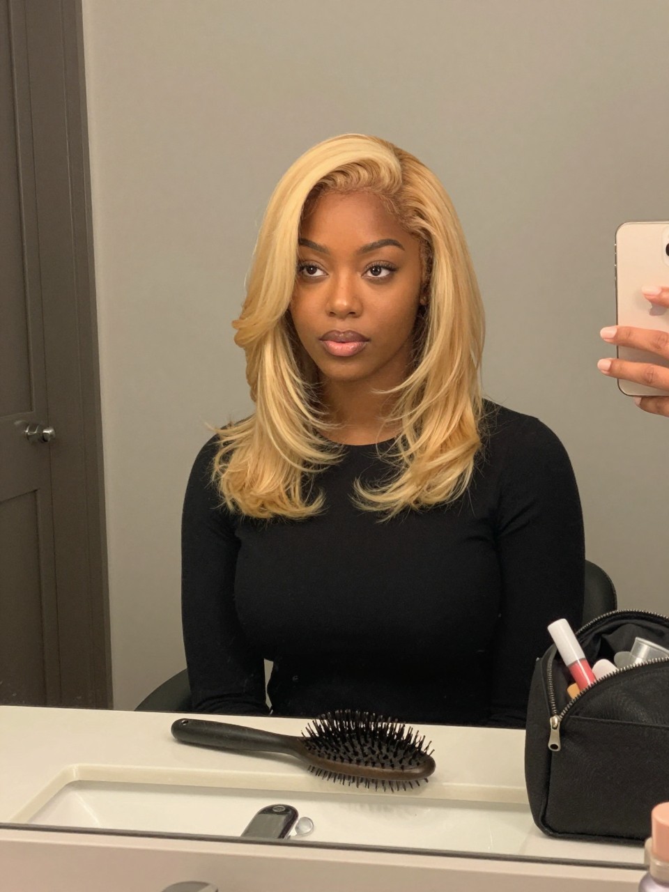 Photo of a black woman wearing butter blonde hair with soft face-framing layers, candid mirror selfie, bathroom vanity mirror setting, soft indoor lighting, with a blowout brush on the counter and a makeup bag nearby, latest iPhone photo quality.