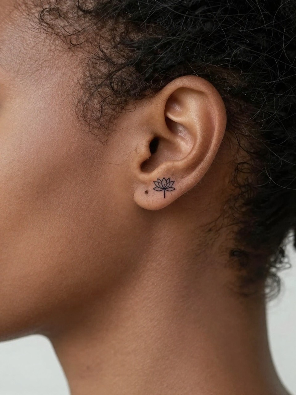 Image Prompt: "Macro shot of a tiny black ink lotus flower tattoo placed behind the ear of a black feminine woman, minimalist fine line design, very small delicate outline, soft clean skin texture, a few loose strands of hair framing the placement, simple feminine styling, tight crop, minimal background, realistic modern tattoo inspiration photo with tattoo as the hero (no watermarks on images)"