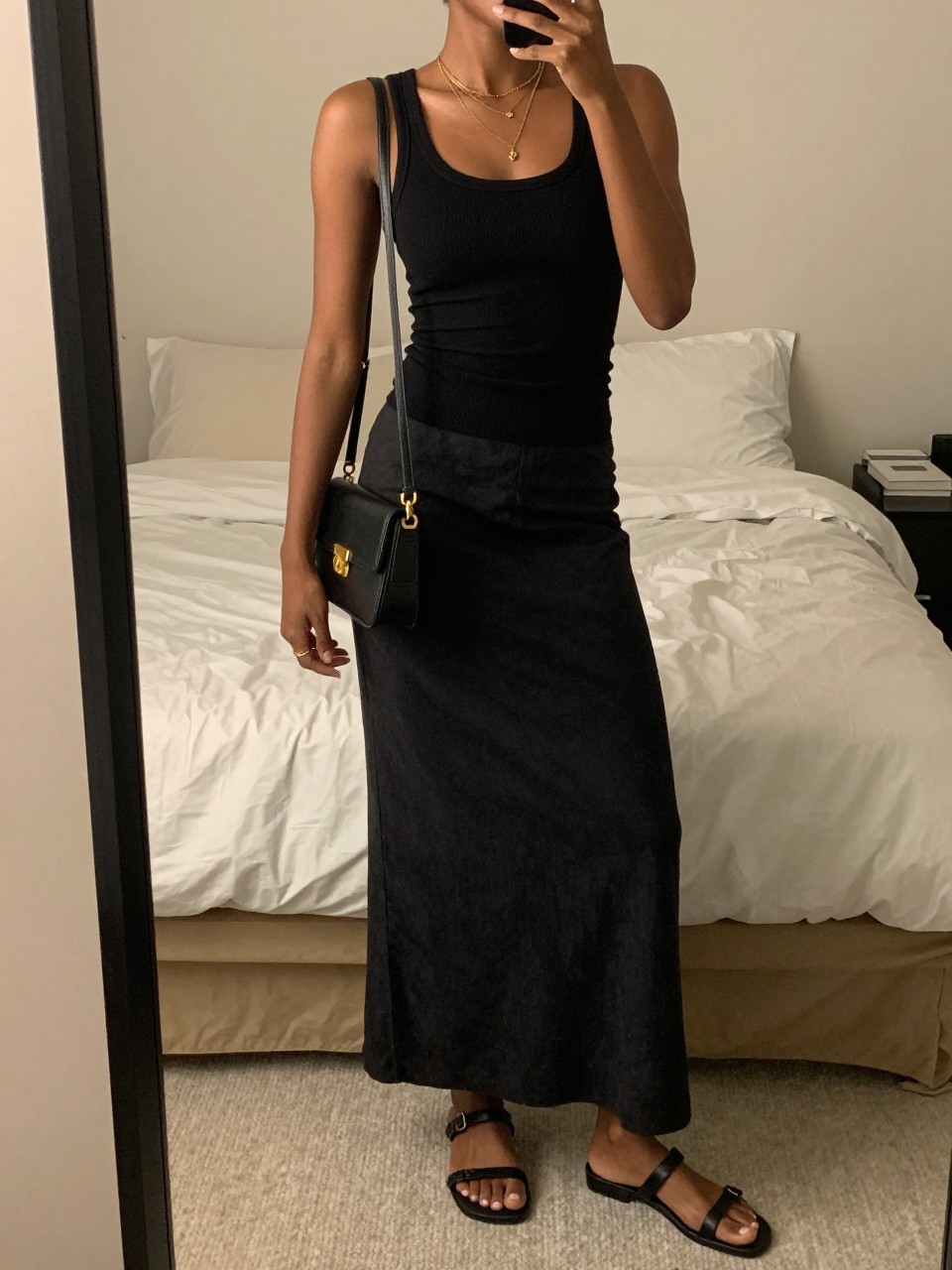mirror selfie of a black woman wearing a black fitted ribbed tank top tucked into a black linen maxi skirt with layered gold necklaces and black flat leather sandals, holding a black crossbody bag with gold hardware, standing in a relaxed minimalist pose, cropped at NECK so face doesn't show, in a feminine-themed bedroom, warm moody lighting, iPhone photo quality.