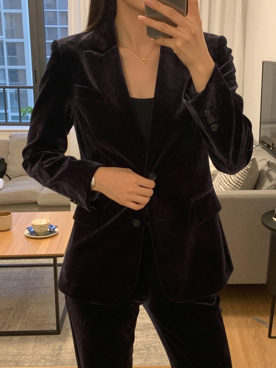 iPhone mirror selfie of a woman in a velvet set, buttoning her blazer, cropped at NECK so face doesn't show, in a city apartment, warm moody lighting, iPhone photo quality.