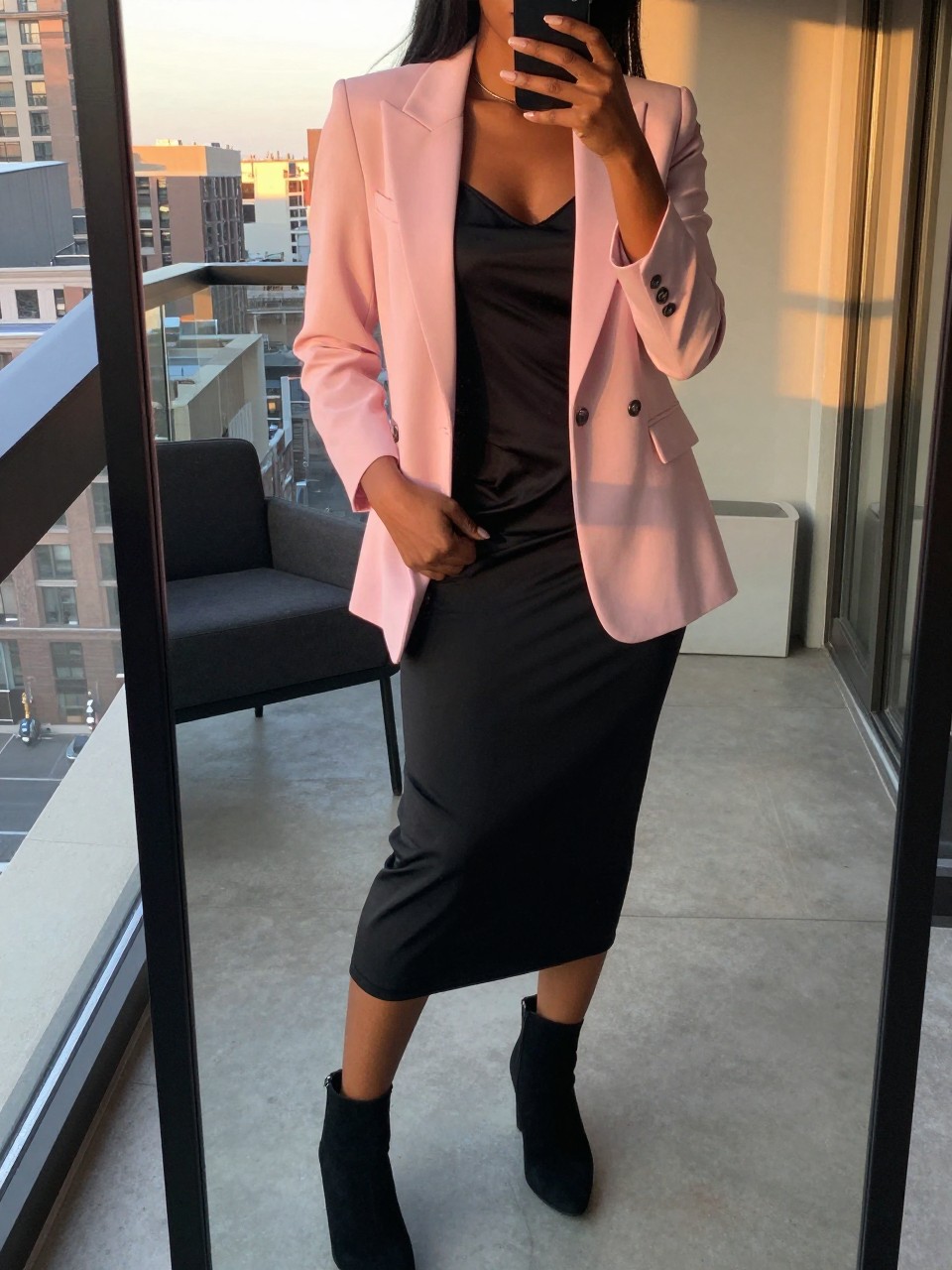 iPhone mirror selfie of a black woman in a pink blazer over a black slip dress with ankle boots, standing with hand in blazer pocket, cropped at NECK so face doesn't show, in a city apartment, evening golden hour, iPhone photo quality.