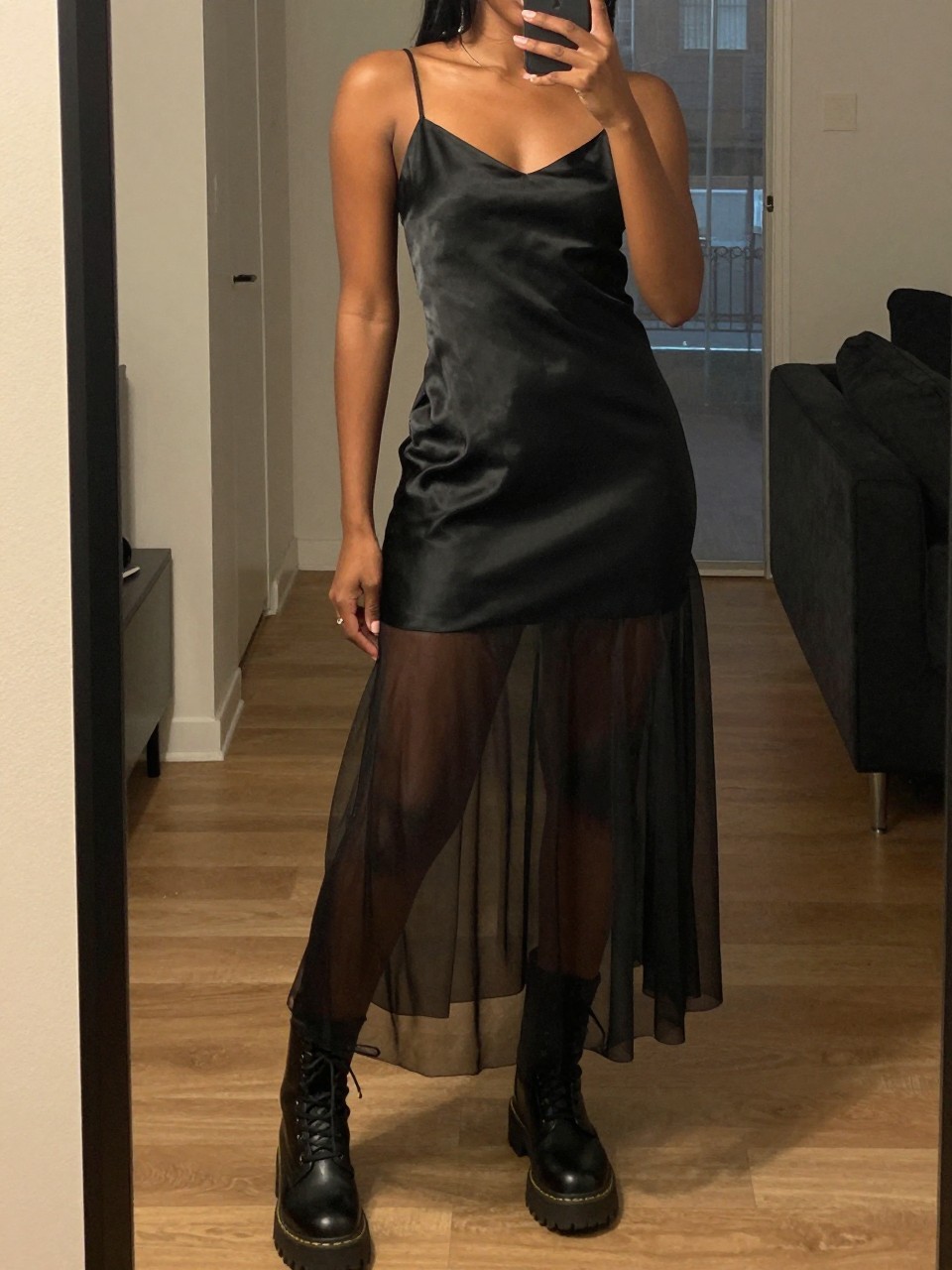 mirror selfie of a black woman wearing a black silk slip dress with a black sheer mesh skirt layered over it and chunky combat boots, one hand holding her phone at her side, cropped at NECK so face doesn't show, in a city apartment, warm moody lighting, iPhone photo quality.