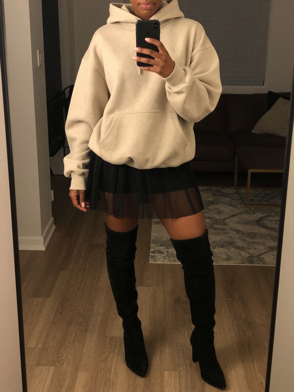 mirror selfie of a black woman wearing an oversized oatmeal hoodie with a black sheer mesh skirt and black suede thigh-high boots, one hand holding her phone at chest level, cropped at NECK so face doesn't show, in a city apartment, warm moody lighting, iPhone photo quality.