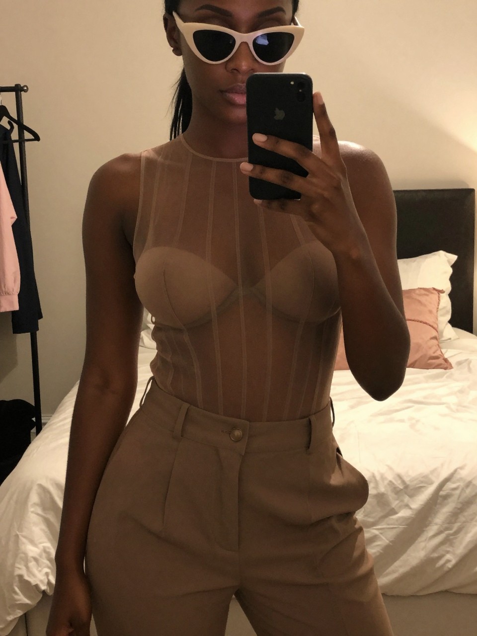 mirror selfie of a black woman in a sheer-paneled top and trousers, wearing cat-eye sunglasses, cropped at NECK so face doesn't show, in a feminine-themed bedroom, warm moody lighting, iPhone photo quality.