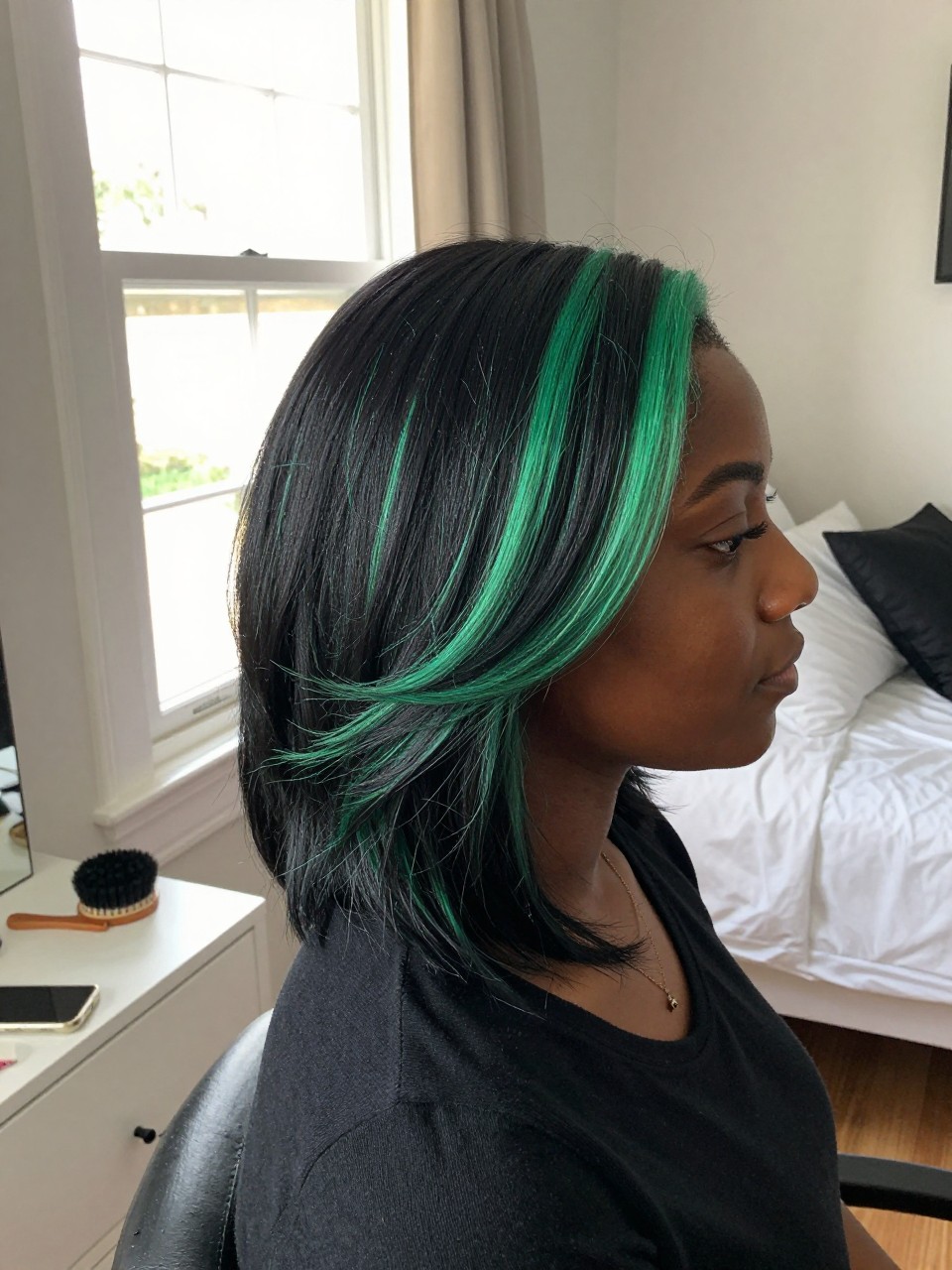 Photo of a black woman wearing a side part black blowout with jade green highlights, side profile view, in a sun-drenched bedroom, natural window light, containing a small lived-in detail such as velcro rollers on the vanity and a round brush nearby, latest iPhone photo quality.