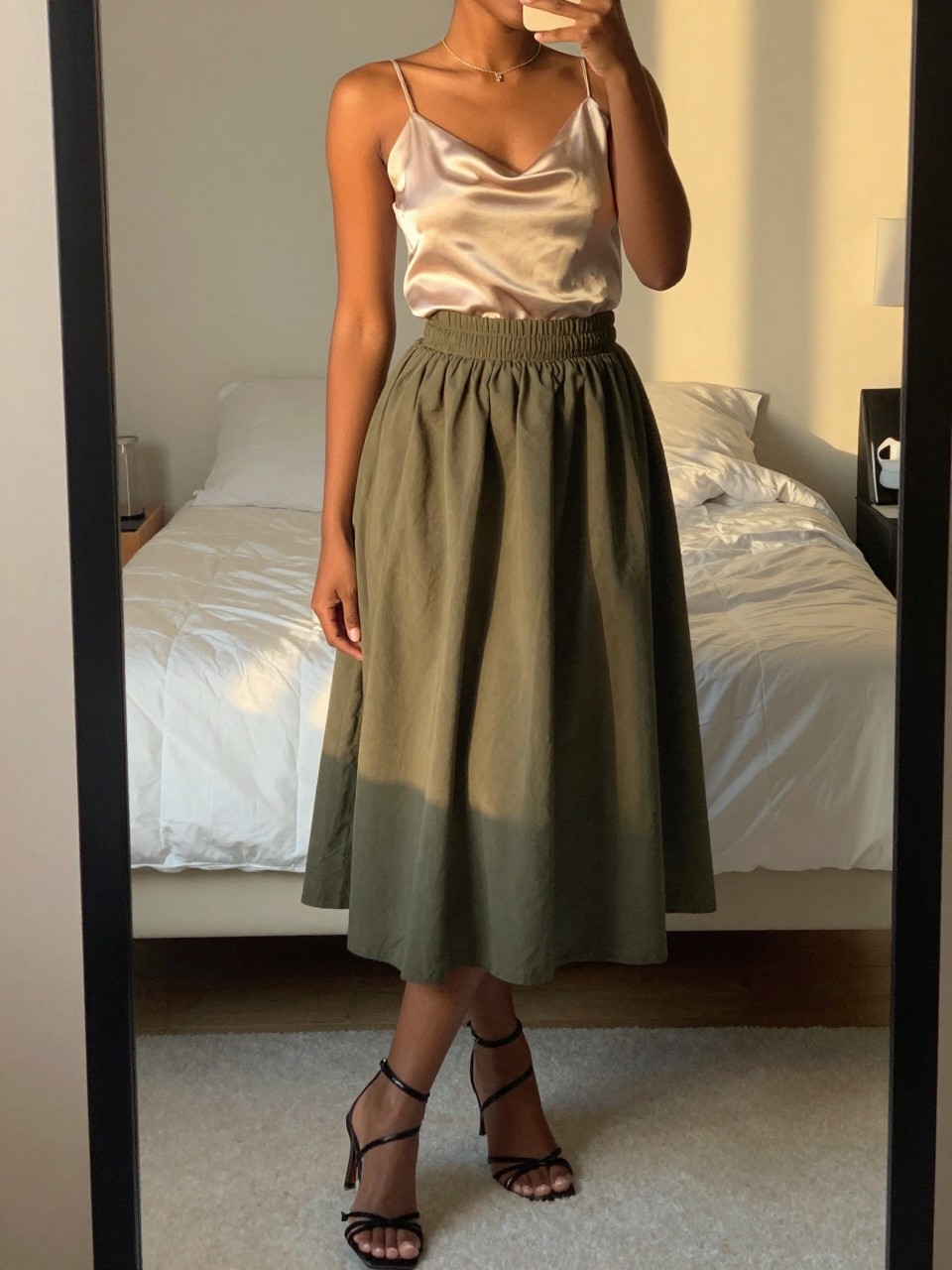 mirror selfie of a black woman in a champagne silk camisole and an olive green balloon skirt with strappy heels, posing with one foot slightly forward, cropped at NECK so face doesn't show, in a feminine-themed bedroom, evening golden hour, iPhone photo quality.