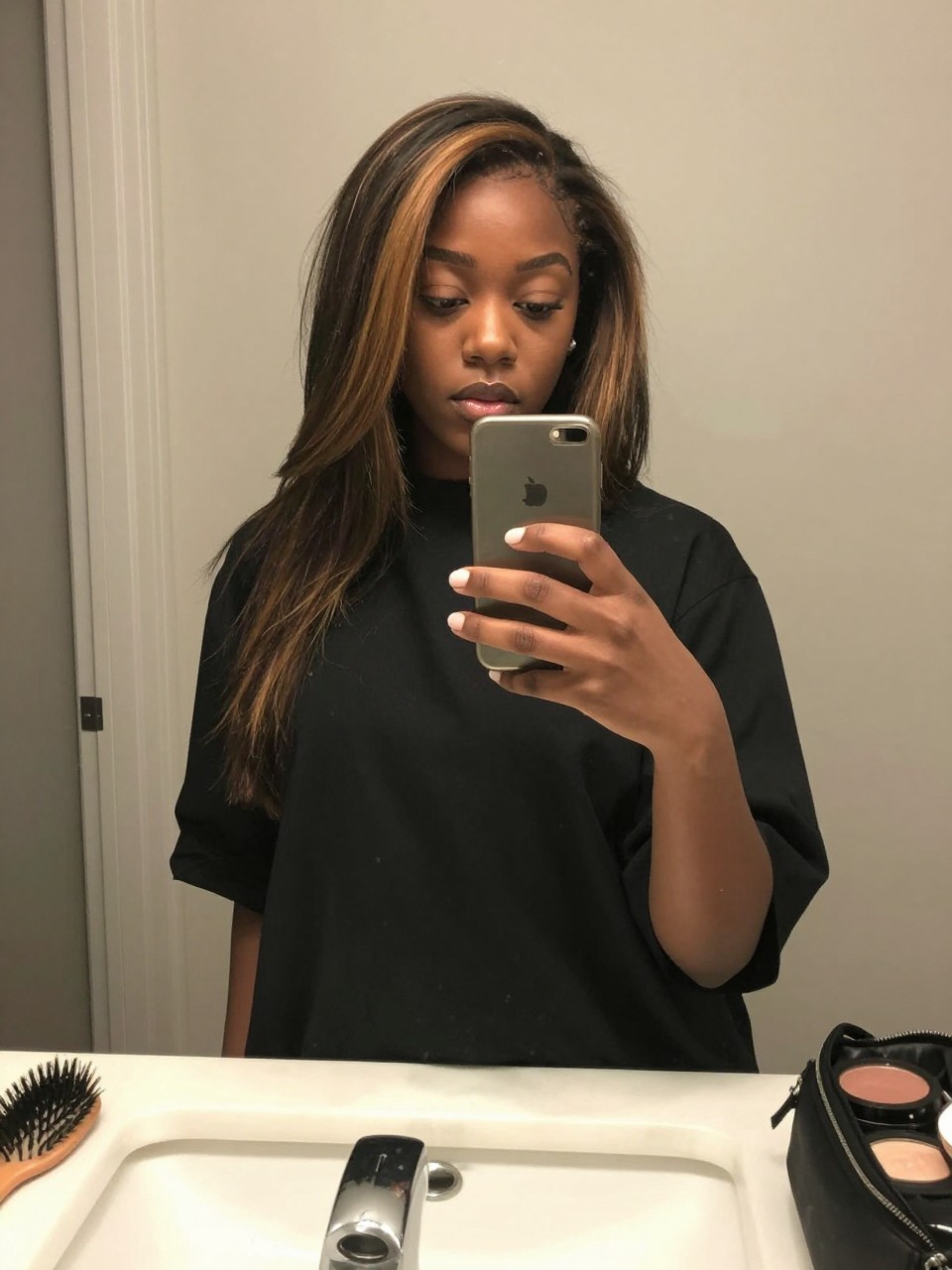 Photo of a black woman wearing a sleek black lob with caramel face-framing highlights, candid mirror selfie, at a bathroom vanity mirror, soft indoor lighting, containing a small lived-in detail such as a hairbrush on the vanity and a makeup bag nearby, latest iPhone photo quality.