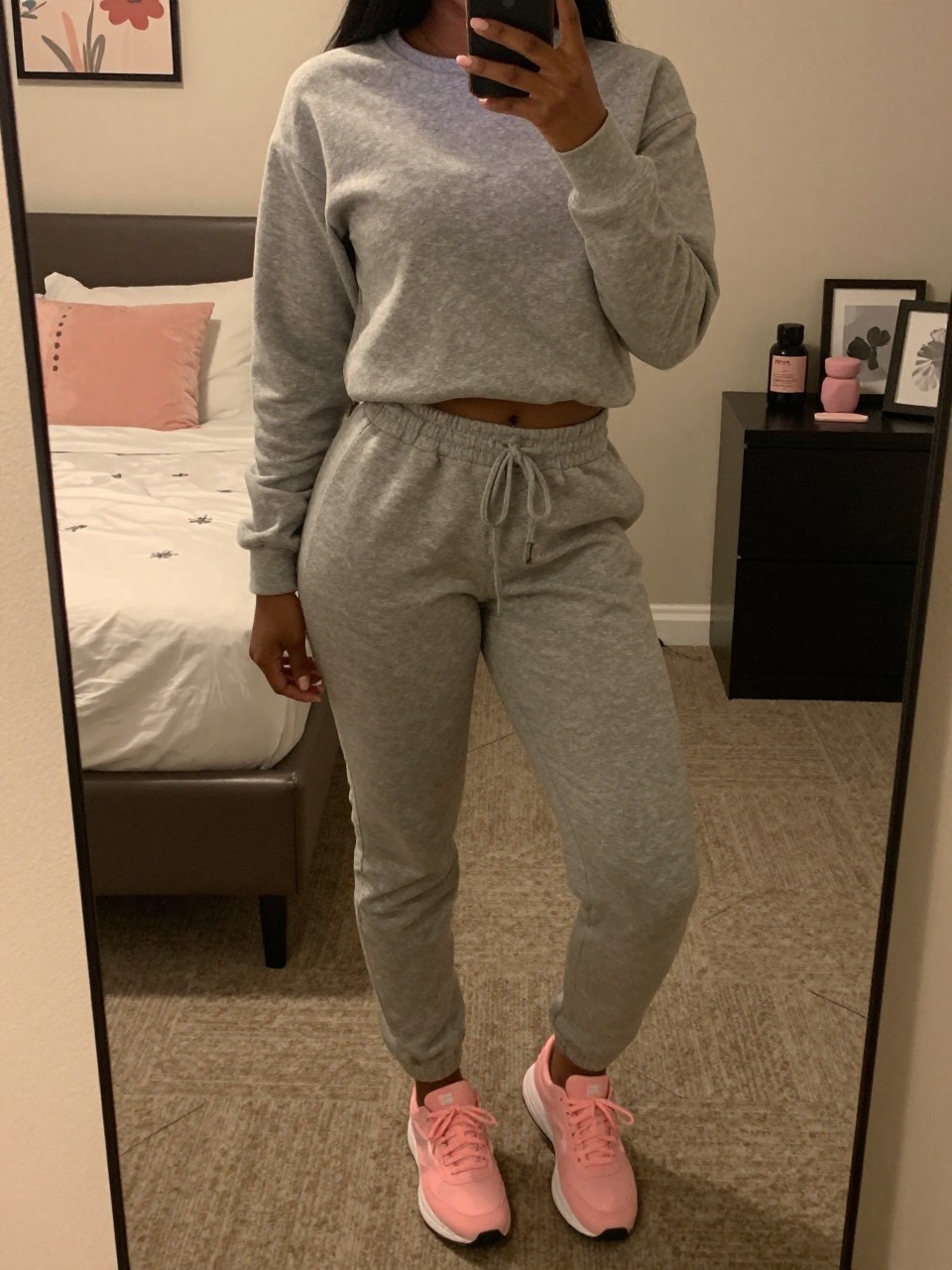 iPhone mirror selfie of a black woman in a grey sweatsuit and pink sneakers, standing with phone near waist, cropped at NECK so face doesn't show, in a feminine-themed bedroom, warm moody lighting, iPhone photo quality.
