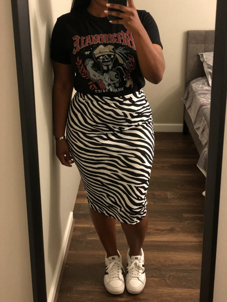 mirror selfie of a curvy black woman in a black graphic tee and a zebra print skirt with white sneakers, casually leaning against a doorframe, cropped at NECK so face doesn't show, in a city apartment, warm moody lighting, iPhone photo quality.