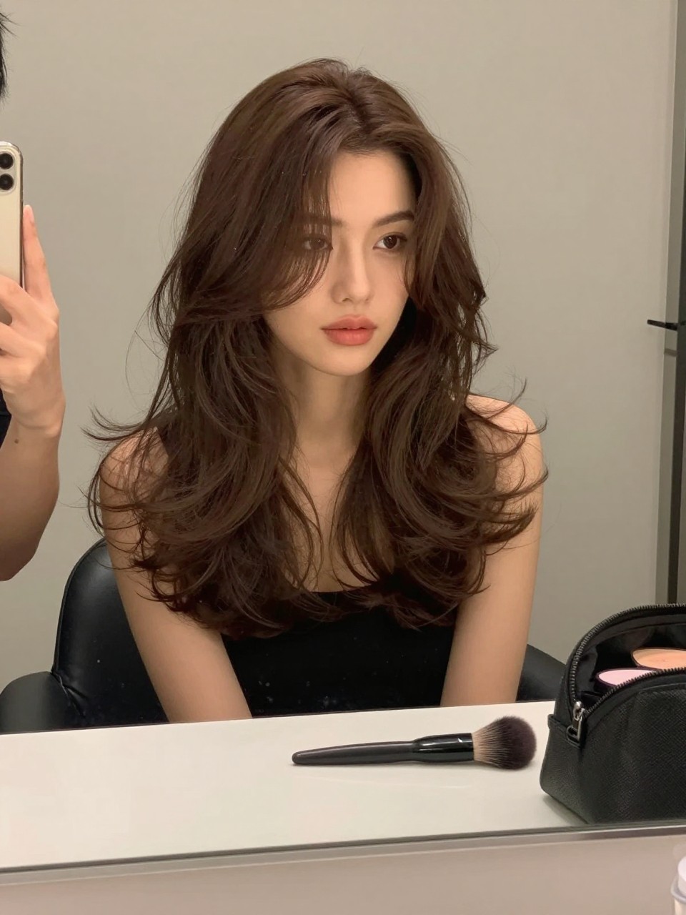 Photo of a woman wearing milk chocolate brown hair in a soft bouncy blowout, candid mirror selfie, bathroom vanity mirror setting, soft indoor lighting, with a round brush on the counter and a makeup bag nearby, latest iPhone photo quality.