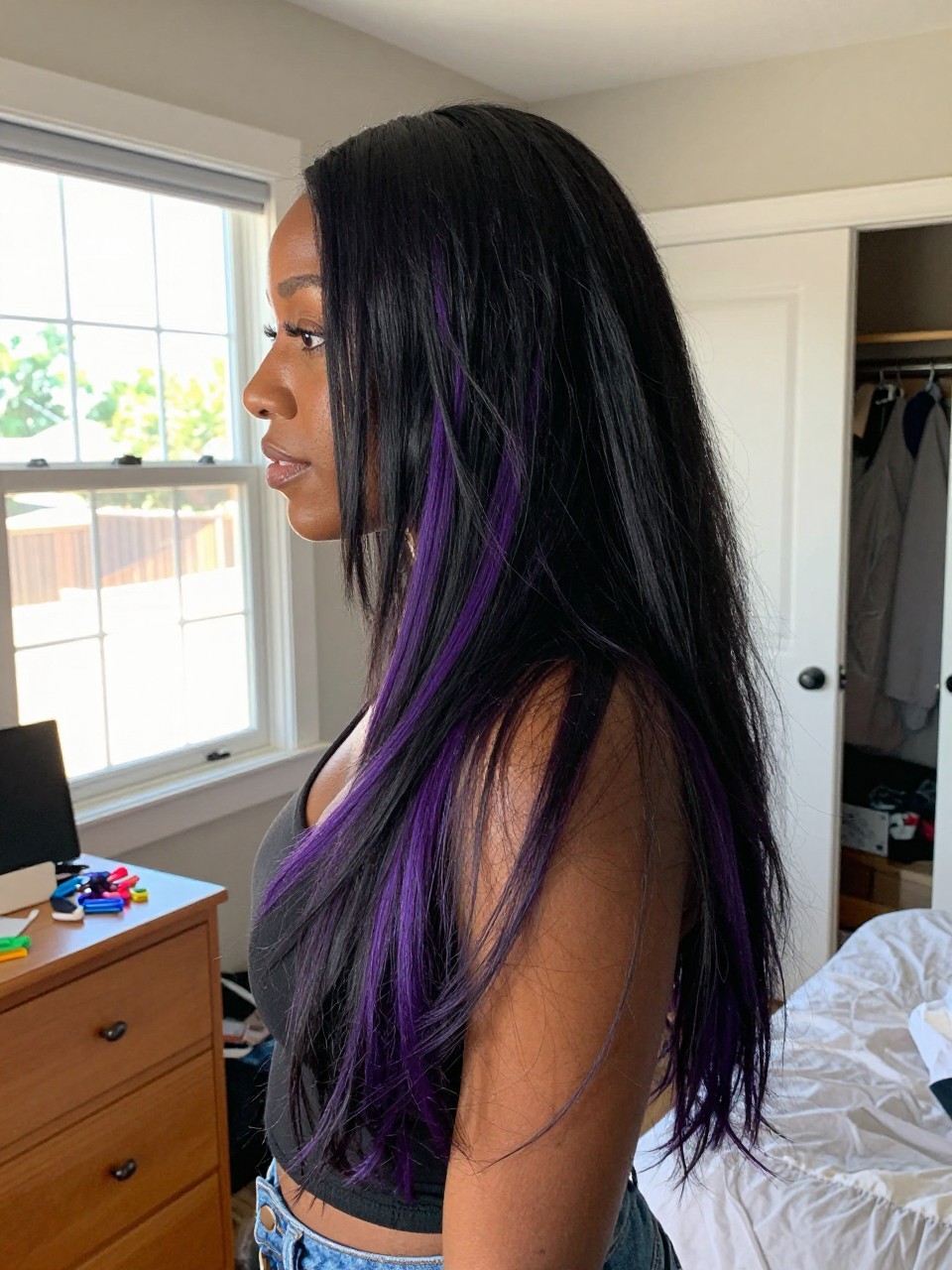 Photo of a black woman wearing long black layered hair with amethyst purple highlights and soft body, side profile view, in a sun-drenched bedroom, natural window light, containing a small lived-in detail such as sectioning clips on the dresser and a slightly open closet behind, latest iPhone photo quality.
