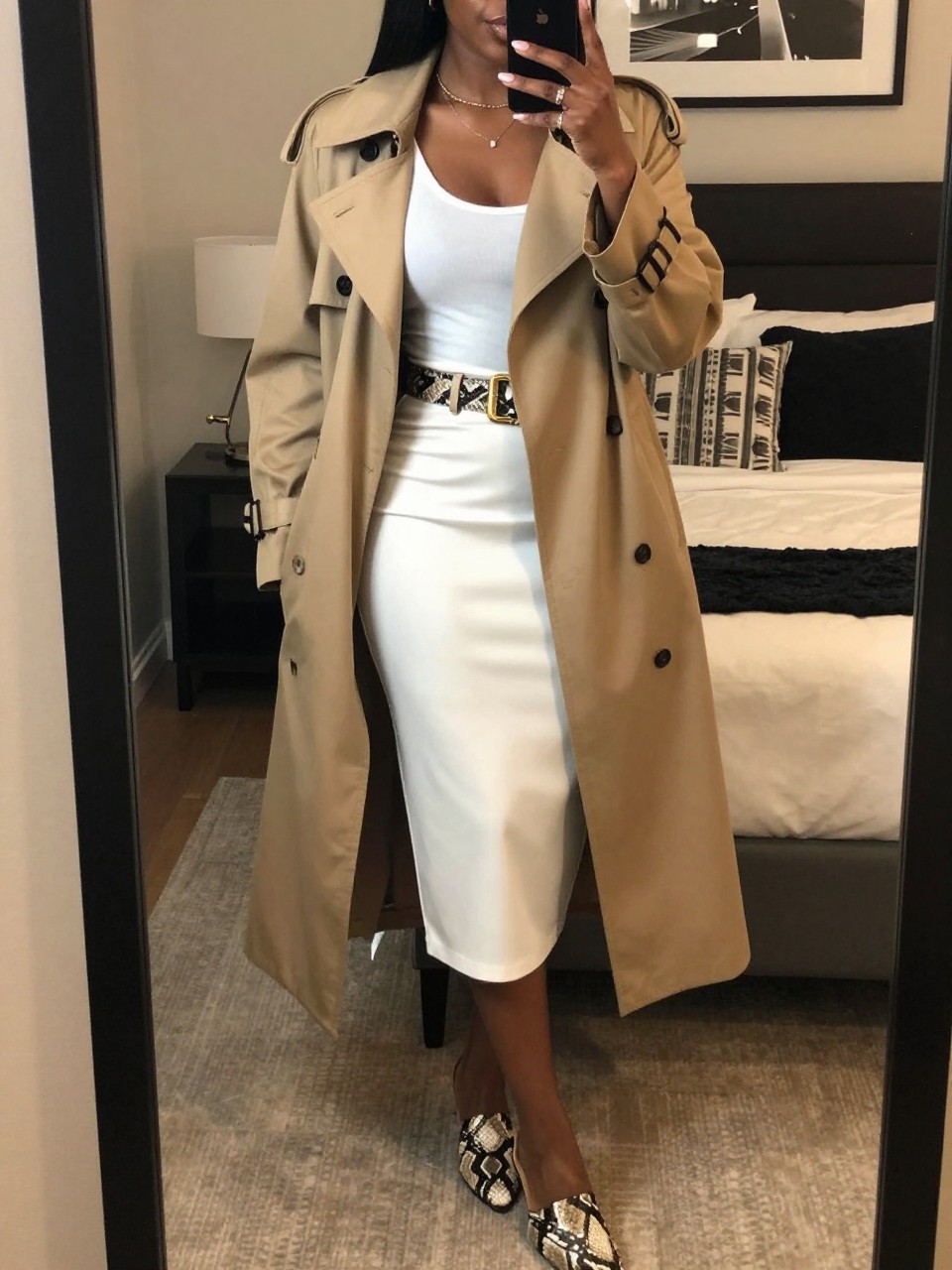 mirror selfie of a black woman wearing a tan trenchcoat open over a white tank top tucked into a white midi skirt, cinched with a snake-print belt, with snake-print mules, holding oversized sunglasses, standing in a sophisticated city pose, cropped at NECK so face doesn't show, in a feminine-themed bedroom, warm moody lighting, iPhone photo quality.