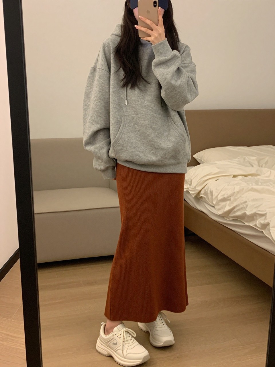 mirror selfie of a woman wearing an oversized heather grey hoodie with a rust brown jersey knit maxi skirt and chunky white sneakers, a baseball cap on, standing in a casual stance, cropped at NECK so face doesn't show, in a feminine-themed bedroom, warm moody lighting, iPhone photo quality.