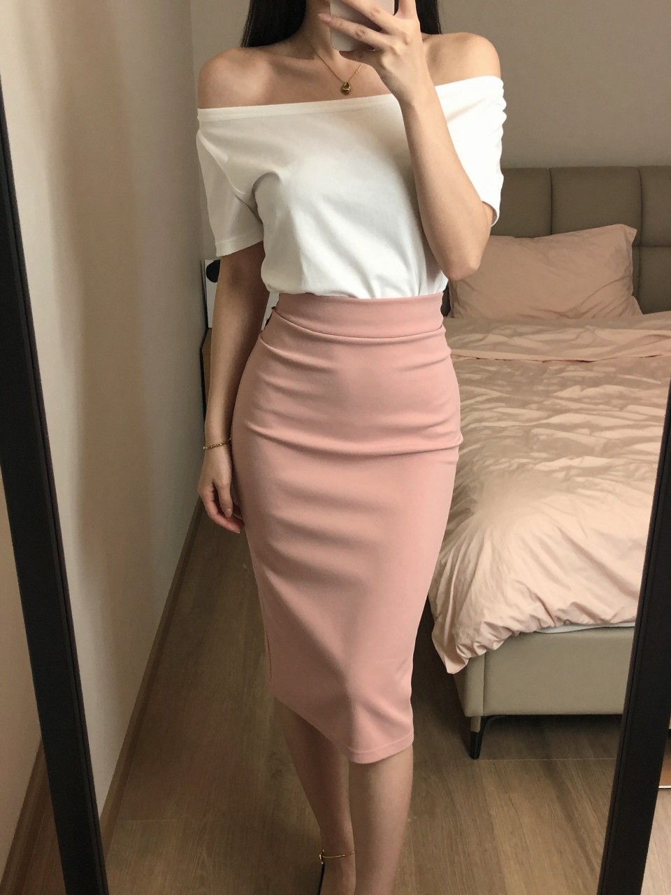 iPhone mirror selfie of a woman wearing an off-the-shoulder white top tucked into a pink pencil skirt with flat sandals, one foot slightly forward showing a gold anklet, cropped at NECK so face doesn't show, in a feminine-themed bedroom, warm moody lighting, iPhone photo quality.