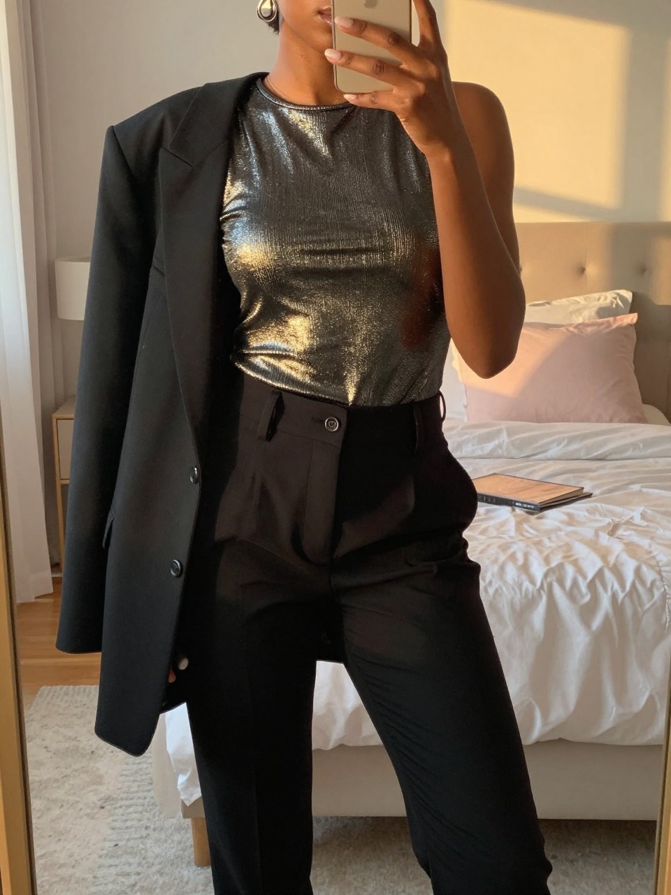 mirror selfie of a black woman in a metallic top and black trousers, blazer over arm, cropped at NECK so face doesn't show, in a feminine-themed bedroom, evening golden hour, iPhone photo quality.