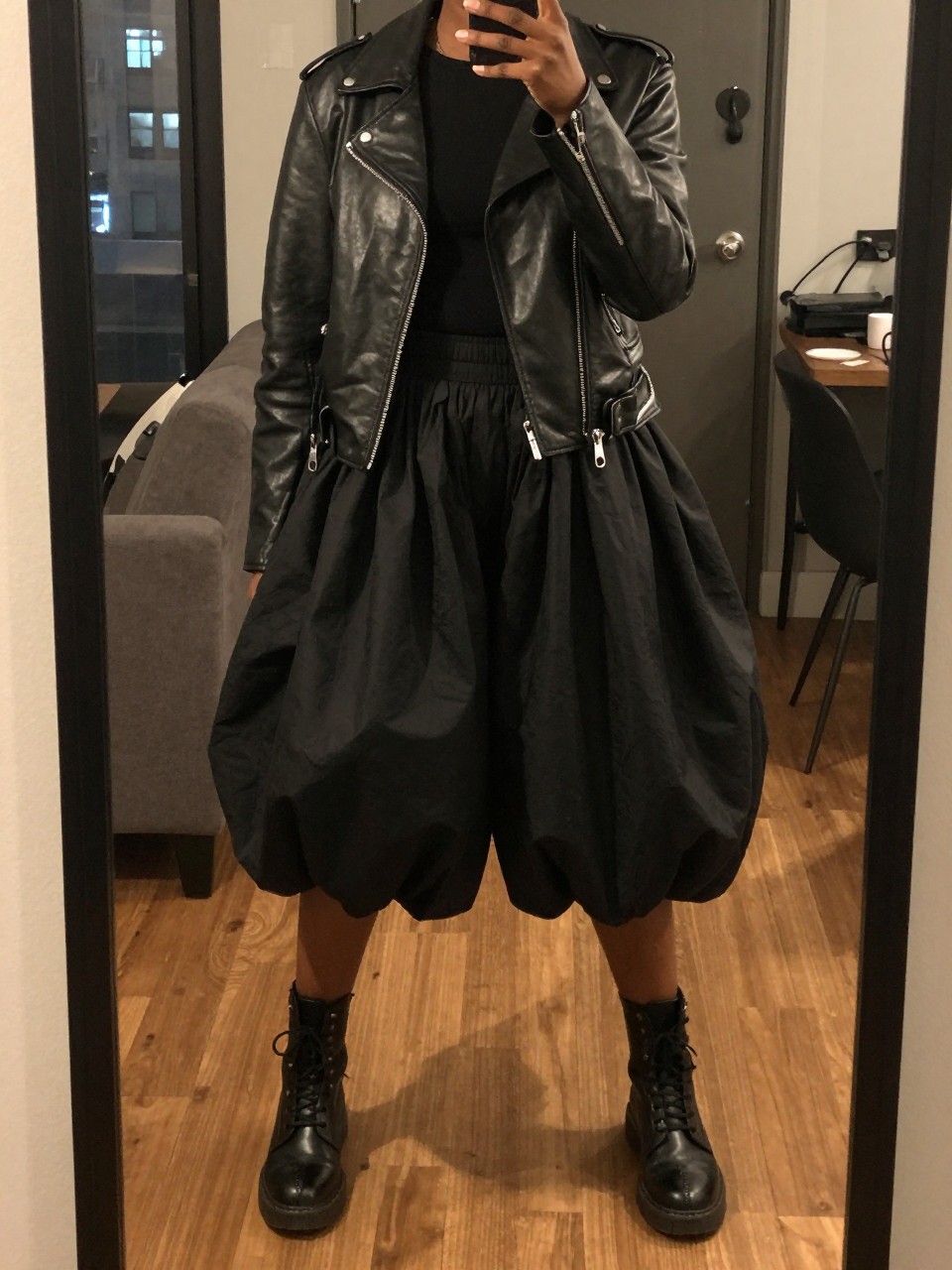 mirror selfie of a black woman in a leather jacket and a black balloon skirt with combat boots, standing with feet shoulder-width apart, cropped at NECK so face doesn't show, in a city apartment, warm moody lighting, iPhone photo quality.
