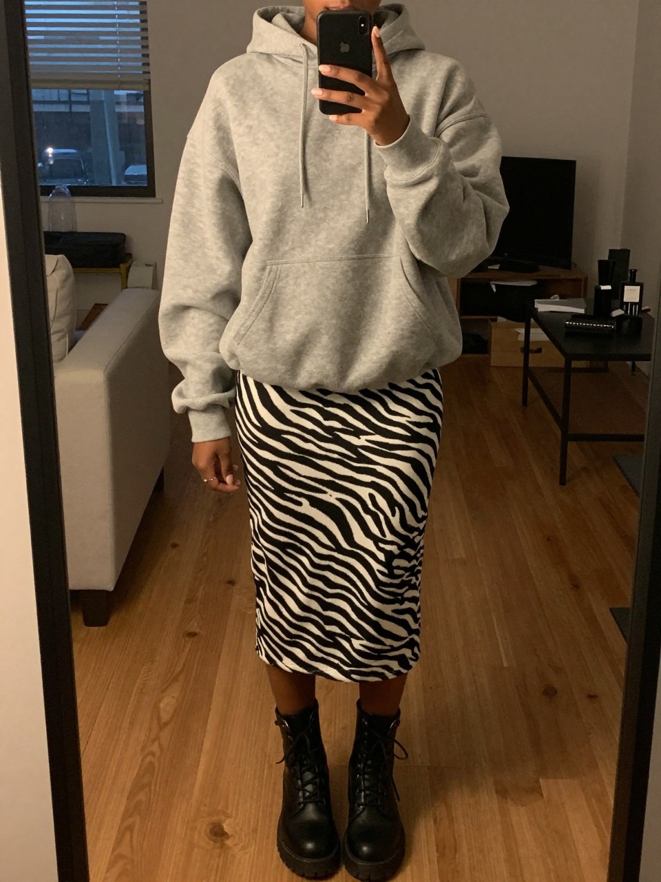 mirror selfie of a black woman in a grey oversized hoodie and a zebra print skirt with combat boots, phone in hand, cropped at NECK so face doesn't show, in a city apartment, warm moody lighting, iPhone photo quality.