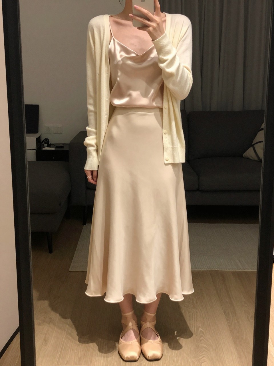 iPhone mirror selfie of a woman wearing a champagne silk camisole under a soft cream cardigan with a matching cream flowy midi skirt and nude ballet flats, one hand holding her phone at chest level, cropped at NECK so face doesn't show, in a city apartment, warm moody lighting, iPhone photo quality.