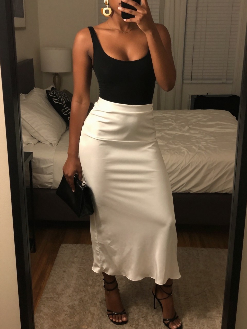 mirror selfie of a black woman wearing a black scoop-neck bodysuit tucked into a white satin maxi skirt with black strappy heeled sandals and bold gold statement earrings, holding a small black clutch, standing in a confident evening pose, cropped at NECK so face doesn't show, in a feminine-themed bedroom, warm moody lighting, iPhone photo quality.