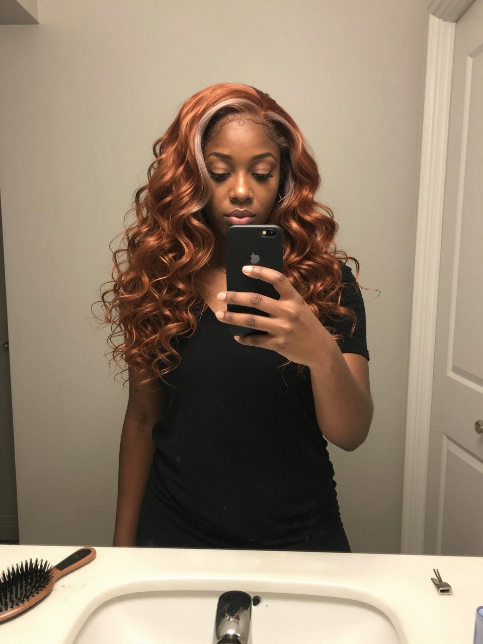 Photo of a black woman wearing a wavy copper lob with ash blonde highlights, candid mirror selfie, at a bathroom vanity mirror, soft indoor lighting, containing a small lived-in detail such as a hairbrush on the sink and a silver clip nearby, latest iPhone photo quality.