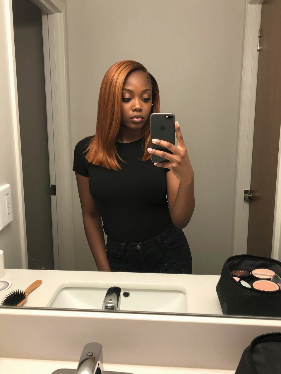 Photo of a black woman wearing a sleek ginger lob with honey highlight ribbons, candid mirror selfie, at a bathroom vanity mirror, soft indoor lighting, containing a small lived-in detail such as a hairbrush on the vanity and a makeup bag nearby, latest iPhone photo quality.