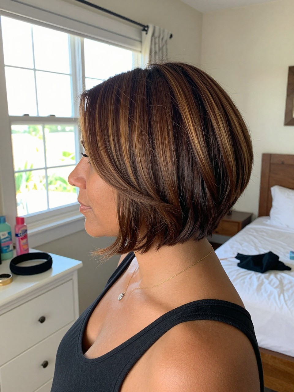 Photo of a carribean woman wearing a short brown French bob with subtle honey highlights and softly curved ends, side profile view, in a sun-drenched bedroom, natural window light, containing a small lived-in detail such as a black headband on the dresser and a hairbrush nearby, latest iPhone photo quality.