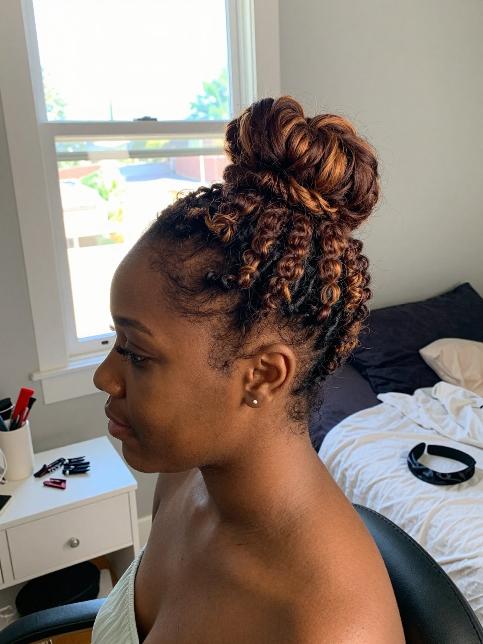 Photo of a Black woman wearing a natural bantu knot-out with cinnamon highlights throughout the springy curls, side profile view, in a sun-drenched bedroom, natural window light, containing a small lived-in detail such as sectioning clips on the vanity and a satin headband nearby, latest iPhone photo quality.