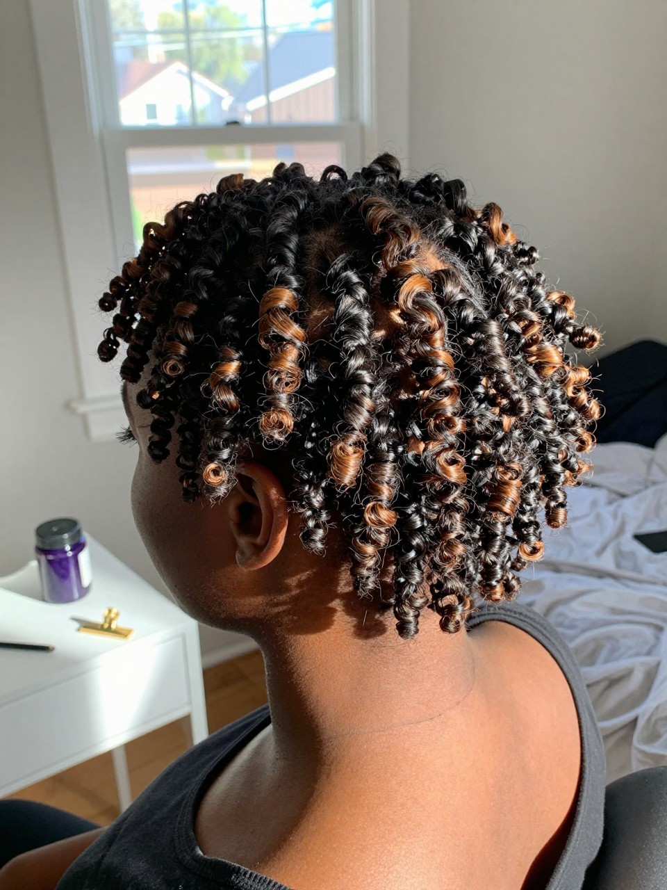 Photo of a Black woman wearing short finger coils with caramel highlights throughout the defined natural curls, back-of-head view to highlight details, in a sun-drenched bedroom, natural window light, containing a small lived-in detail such as a gel jar on the vanity and a gold clip nearby, latest iPhone photo quality.