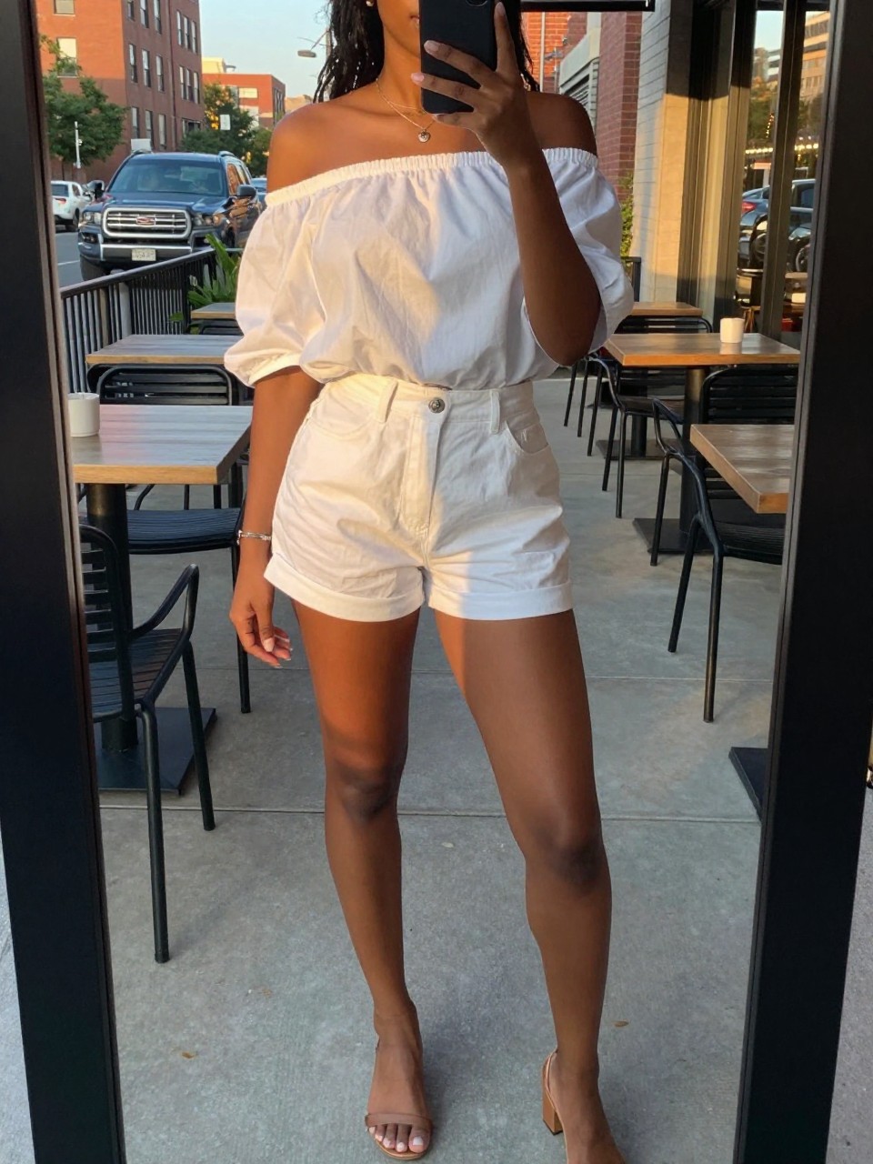 mirror selfie of a black woman in a white off-shoulder top and white shorts with sandals, standing by an outdoor cafe, cropped at NECK so face doesn't show, in a city apartment, evening golden hour, iPhone photo quality.