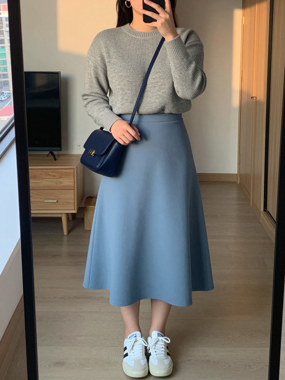 mirror selfie of a curvy woman wearing a heather grey fine-knit sweater tucked into a dusty blue A-line midi skirt with white sneakers and silver hoop earrings, holding a navy crossbody bag, standing in a relaxed elegant pose, cropped at NECK so face doesn't show, in a city apartment, evening golden hour, iPhone photo quality.