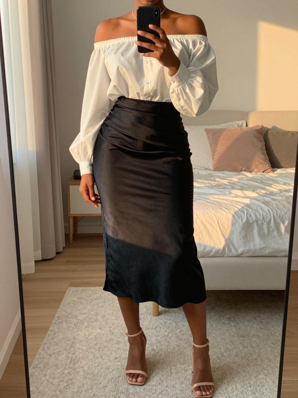 mirror selfie of a black woman wearing a white off-the-shoulder blouse tucked into a black satin midi skirt with nude strappy block-heel sandals, one hand holding her phone at chest level, cropped at NECK so face doesn't show, in a feminine-themed bedroom, evening golden hour, iPhone photo quality.