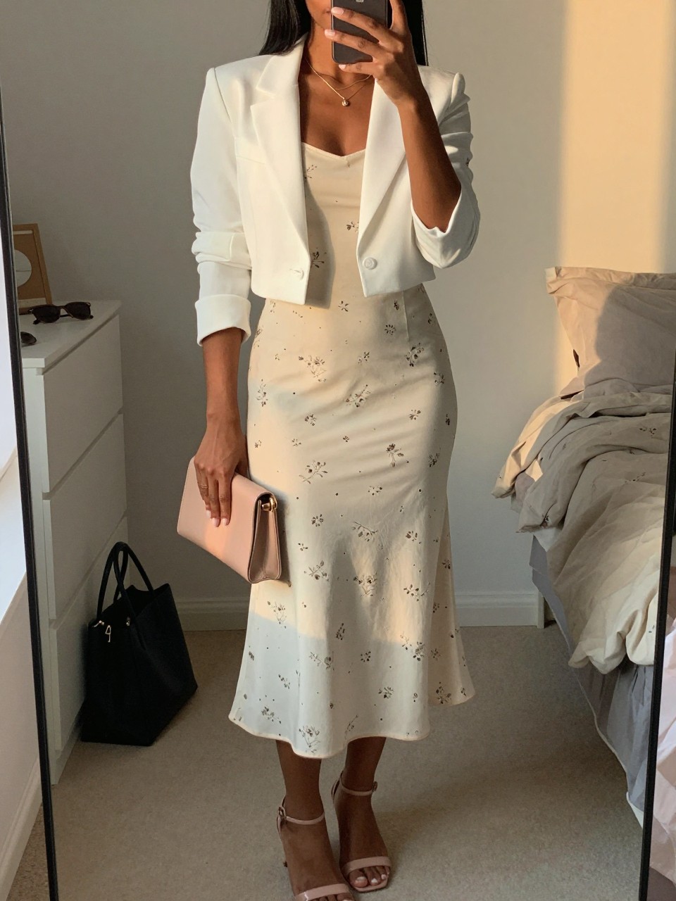 mirror selfie of a black woman wearing a cream printed midi dress with small floral details and a white tailored cropped blazer, nude heeled sandals, a blush mini clutch, and a delicate gold necklace, holding a phone in front of a mirror, cropped at NECK so face doesn't show, with a natural casual selfie pose, in a feminine-themed bedroom, evening golden hour, including small lived-in details like a tote bag on the floor, sunglasses on a dresser, or soft bedding in the background, latest iPhone photo quality.