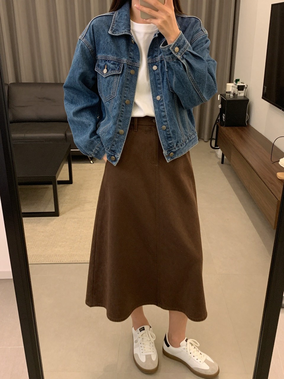 iPhone mirror selfie of a woman wearing a blue denim jacket over a white tee tucked into a chocolate brown cotton A-line skirt with white sneakers, one hand in her jacket pocket, cropped at NECK so face doesn't show, in a city apartment, warm moody lighting, iPhone photo quality.