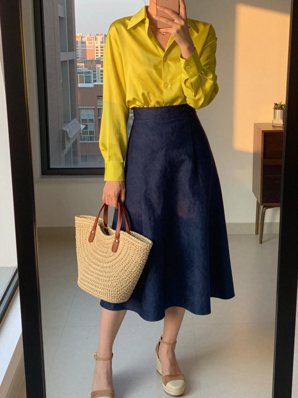 iPhone mirror selfie of a woman wearing a bright yellow silk blouse tucked into a navy linen A-line skirt with a raffia tote and espadrille wedges, one hand holding her phone, cropped at NECK so face doesn't show, in a city apartment, evening golden hour, iPhone photo quality.