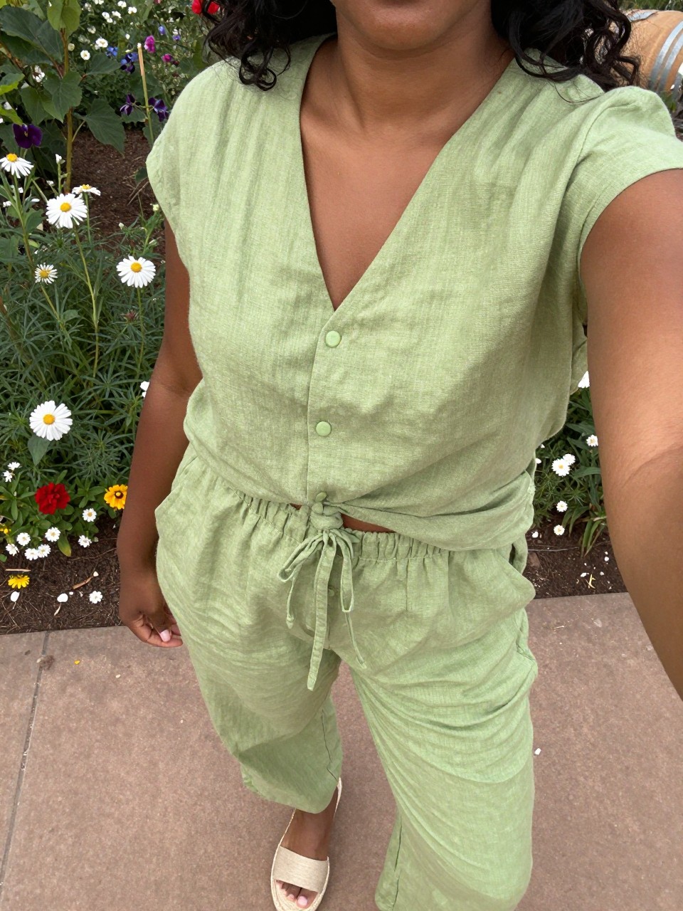 selfie of a black curvy woman in a pistachio green linen set and espadrilles, standing in a garden with flowers, cropped at NECK so face doesn't show, at a winery, warm moody lighting, iPhone photo quality.
