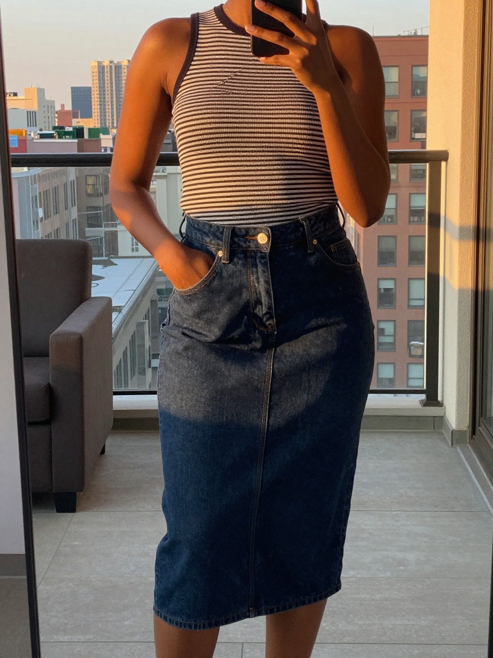 mirror selfie of a Black woman wearing a dark wash denim skirt with a striped top, standing with one hand in her pocket and phone in the other, cropped at NECK so face doesn't show, in a city apartment, evening golden hour light, iPhone photo quality.