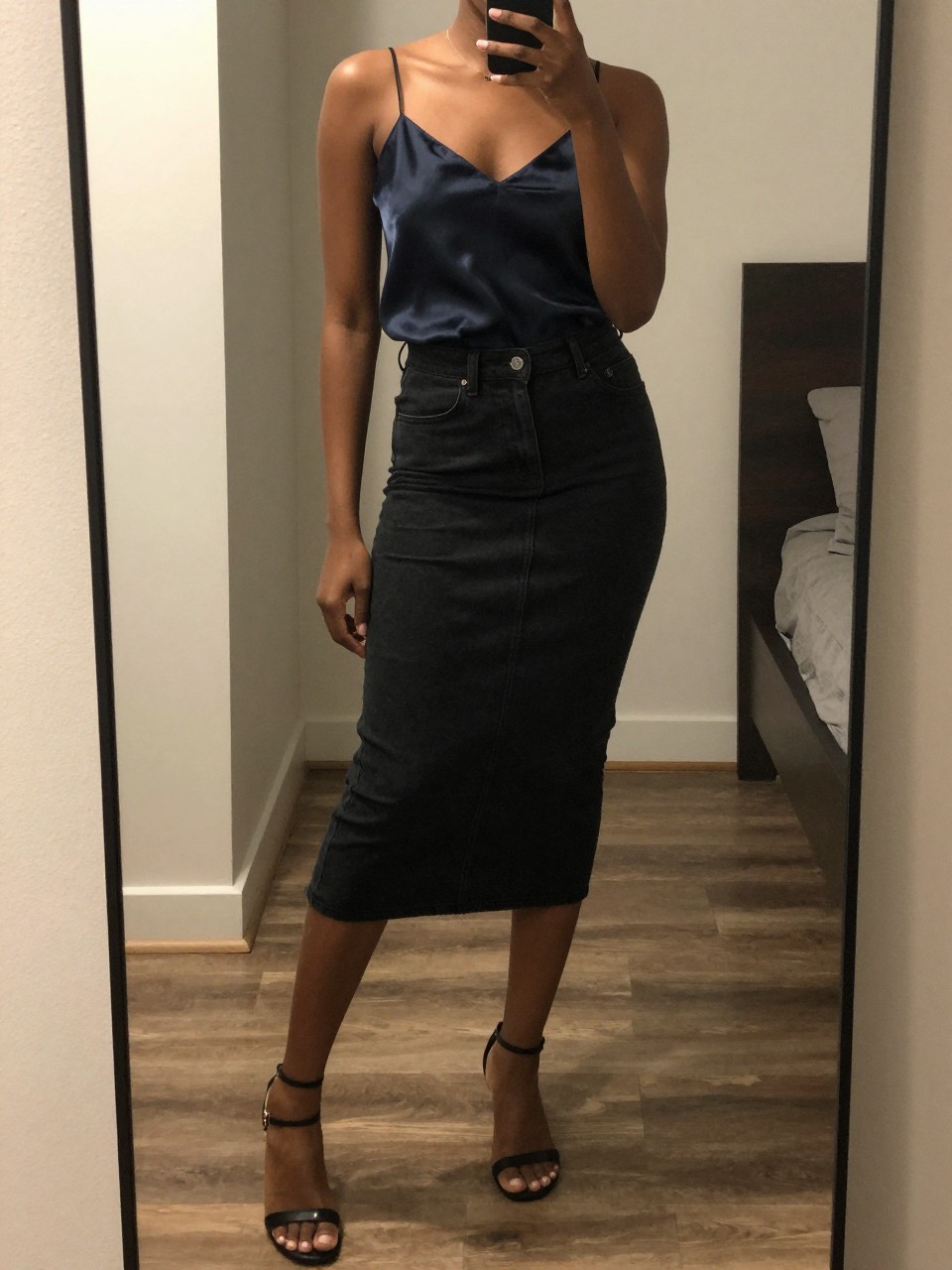 mirror selfie of a Black woman wearing a black denim skirt with a silk cami and heeled sandals, standing with one foot forward and phone in hand, cropped at NECK so face doesn't show, in a city apartment, warm moody lighting, iPhone photo quality.