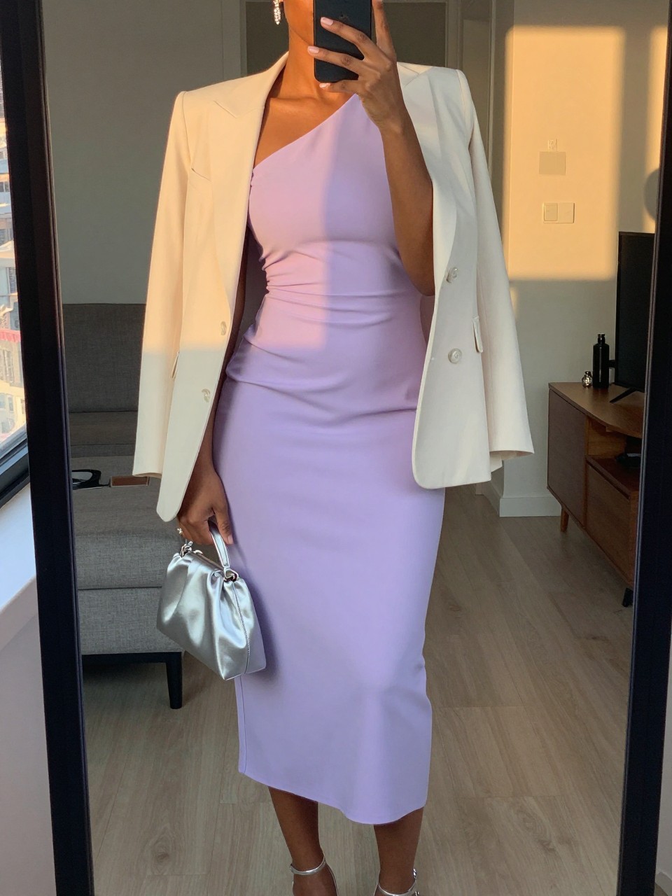 mirror selfie of a black woman wearing a soft lavender crepe one-shoulder midi dress with a cream light blazer, silver strappy heels, simple crystal drop earrings, and a silver satin small clutch, holding a phone in front of a mirror, cropped at NECK so face doesn't show, with a natural casual selfie pose, in a city apartment, evening golden hour, including small lived-in details, latest iPhone photo quality.