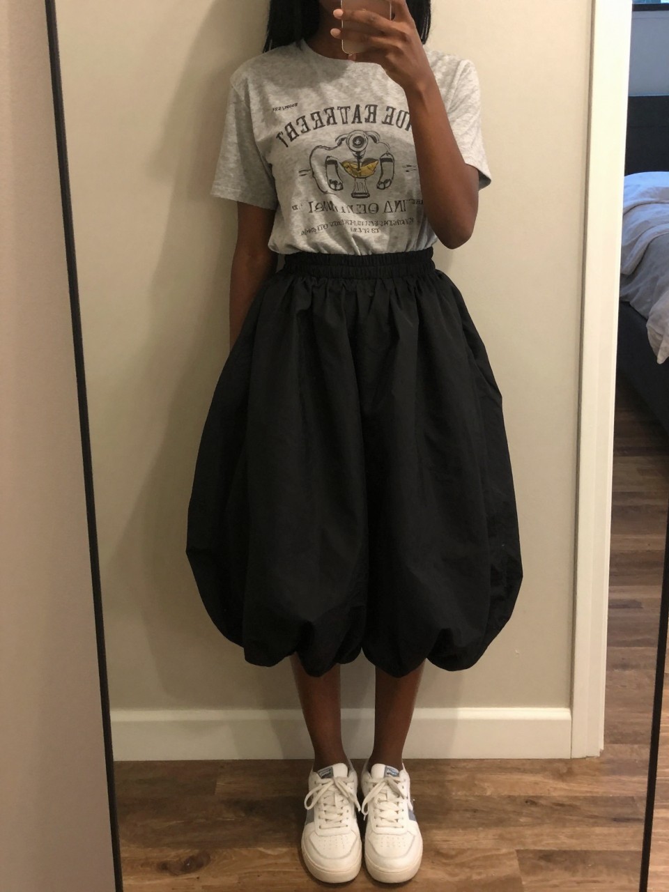 mirror selfie of a black woman in a grey graphic tee and a black balloon skirt with white sneakers, leaning casually against a wall, cropped at NECK so face doesn't show, in a city apartment, warm moody lighting, iPhone photo quality.