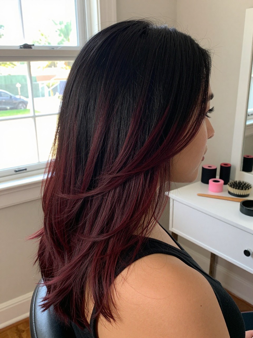 Photo of a woman wearing a side part black blowout with deep burgundy highlights, side profile view, in a sun-drenched bedroom, natural window light, containing a small lived-in detail such as velcro rollers on the vanity and a round brush nearby, latest iPhone photo quality.