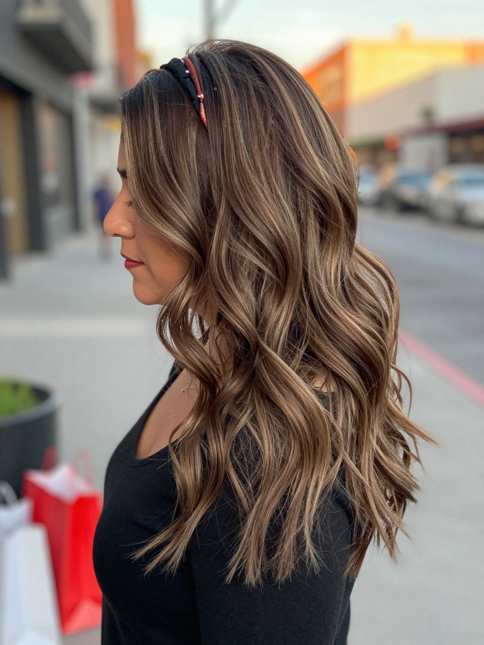 Photo of a mexican woman wearing beachy ash brown waves with dusty highlight streaks and soft texture, side profile view, in a softly blurred city street background, golden hour glow, containing a small lived-in detail such as a headband pushed back and a few shopping bags nearby, latest iPhone photo quality.
