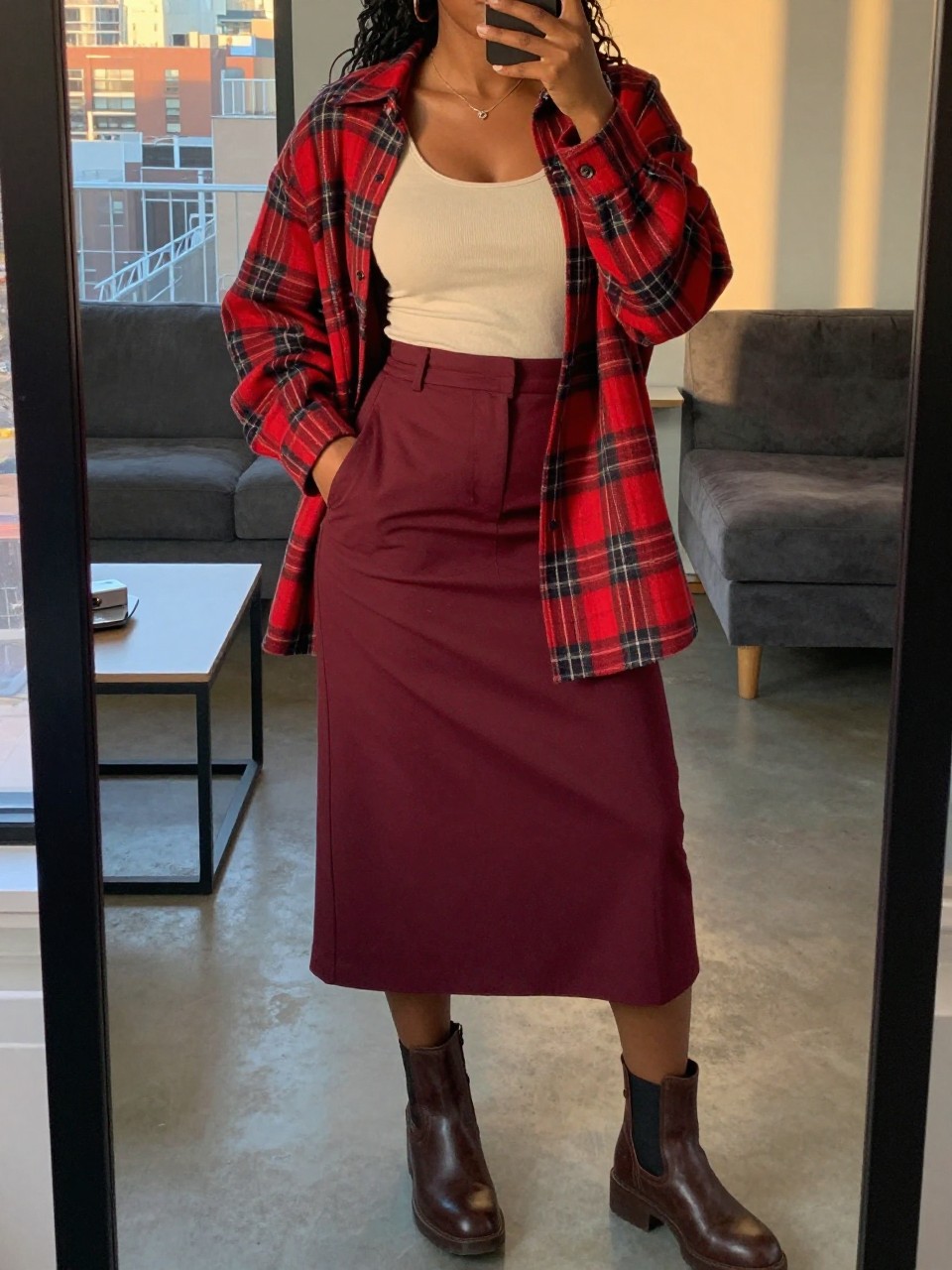 mirror selfie of a black woman wearing an oversized red plaid flannel open over a cream tank top with a deep burgundy A-line skirt and brown chunky ankle boots, one hand in her pocket, cropped at NECK so face doesn't show, in a city apartment, evening golden hour, iPhone photo quality.