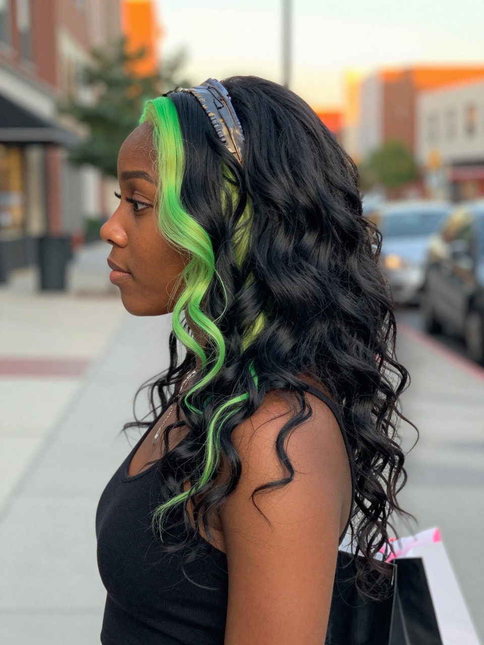 Photo of a black woman wearing beachy black waves with neon green highlight streaks and soft texture, side profile view, in a softly blurred city street background, golden hour glow, containing a small lived-in detail such as a headband pushed back and a few shopping bags nearby, latest iPhone photo quality.